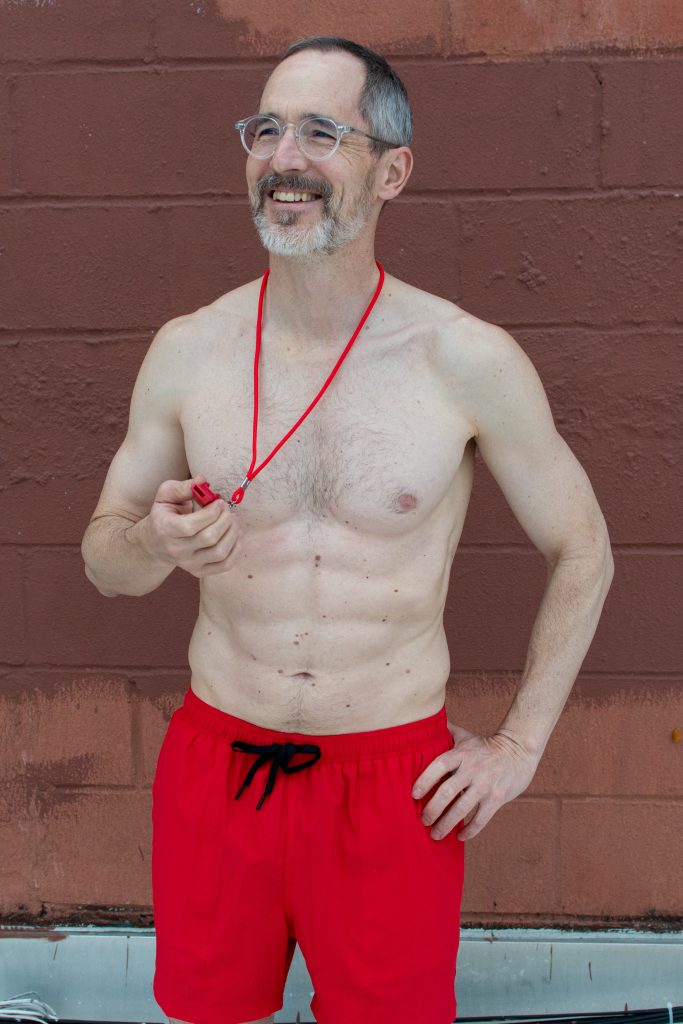 A shirtless middle-aged novelist in a bright red swimsuit, holding a bright red whistle, as he pretends it is not late October. 